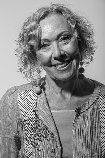 A black and white image of an older woman standing against a plain light grey wall. She is wearing a three piece outfit, dangling ear rings and has a very big smile.