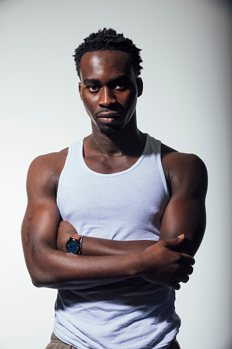 A handsome muscular black man stands against a plain background staring intently at the camera. His arms are folded. He is wearing a plain vest. He has a watch on his wrist.