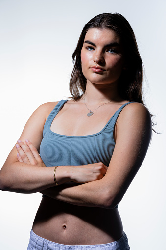 A young woman in a blue crop top and jeans stands against a plain background. Around her neck she has a heart shaped pendant and a gold bangle on her wrist. Her arms are folded and she is staring directly at the camera unsmiling.