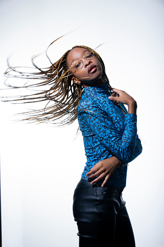 A young black girl wearing glasses a blue long sleeve top and faux leather trousers is standing against a plain back ground.It is a three quarter length image of her standing side on with her arms wrapped around herself. Her braids are flying out behind her in a whipped motion.