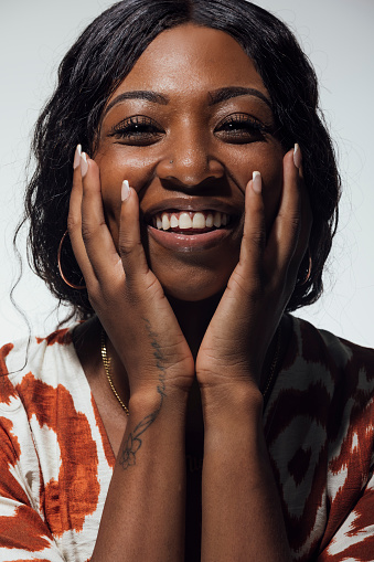 A beautiful black woman is laughing at the camera though her hands which are placed on her cheeks. She is against a light background and is wearing a white and red patterned top. On her right wrist and hand is a small illegible tattoo .