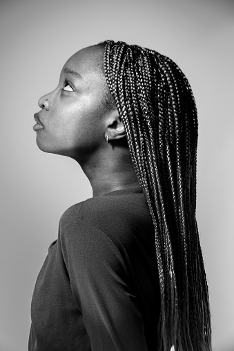 A black and white portrait of a slim black woman wearing a dark top and long braided hair.She is turned away from the camera and is looking up with her braids flowing down her back. The background is grey.