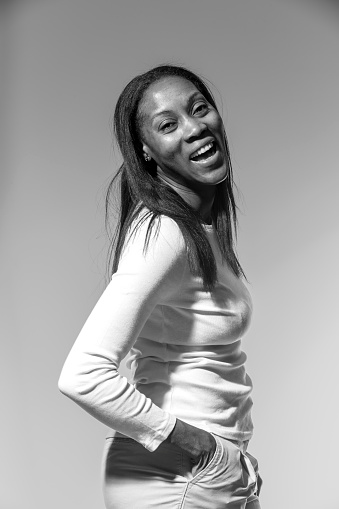 A monochrome image of a 40 something black woman turning to laugh at the camera. She is on a grey background and is wearing trousers and a long sleeved white top.  Her hands are in her pockets. Her hair is black, straight and long.