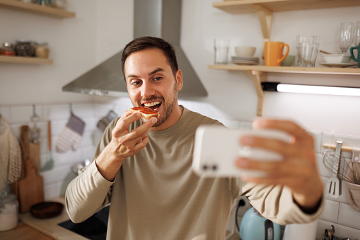 Young man taking selfie while eating toast bread in a domestic kitchen