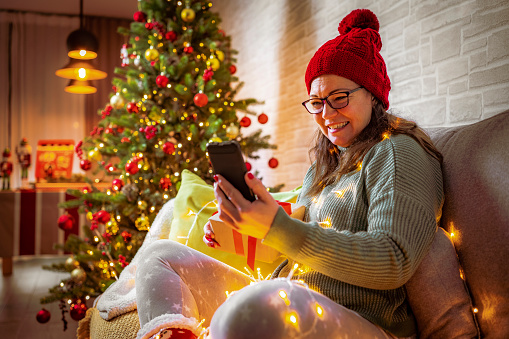 Woman wearing knit hat sitting on sofa using smartphone for video calls with family or friends during Christmas Holidays. Defocused Christmas tree and string lights complete the composition. High resolution 42Mp indoors digital capture taken with Sony A7rII and and Zeiss Batis 40mm F2.0 CF lens