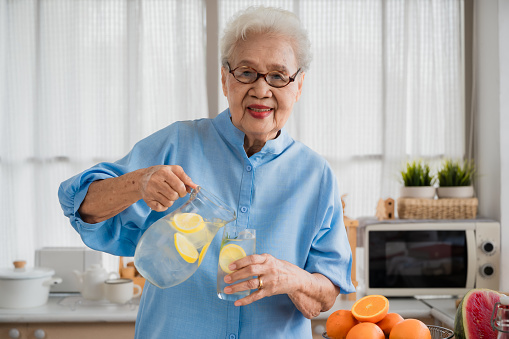 Portrait of happy Asian senior old elderly woman with gray hair wearing glasses standing smiling and pouring iced lemonade from a jar into a glass with looking at camera in the kitchen room at home.