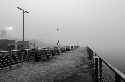 Fisherman's deck on a foggy morning. Concrete structure with stainless steel fences with wooden benches and light poles. Black and white photo.