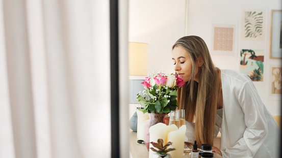 Smelling flowers, beautiful woman and beauty routine of a happy bride wearing a bathrobe while getting ready for her wedding day. Young female enjoying the aroma of fresh roses in her bedroom