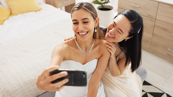 Bride, bridesmaid and selfie with phone, wedding and ring with a smile to post on social media. Happy woman and asian friend excited for marriage celebration with fashion white dress in a bedroom