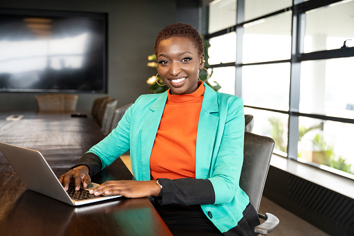 Front view of corporate professional in vibrant attire sitting at conference table and pausing from typing to smile at camera.