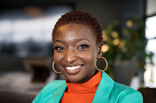 Close view of shorthaired businesswoman wearing teal jacket over orange top, hoop earrings, sitting in modern office and smiling at camera.