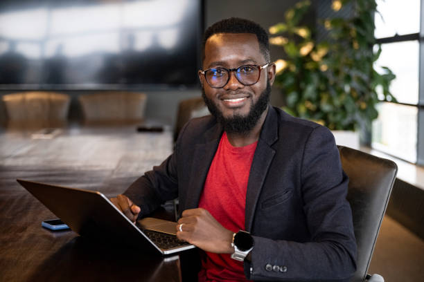 Portrait of bearded African businessman with laptop Corporate professional in early 30s wearing eyeglasses, blazer over red t-shirt, sitting at conference table and pausing from typing to smile at camera. kenyan man stock pictures, royalty-free photos & images