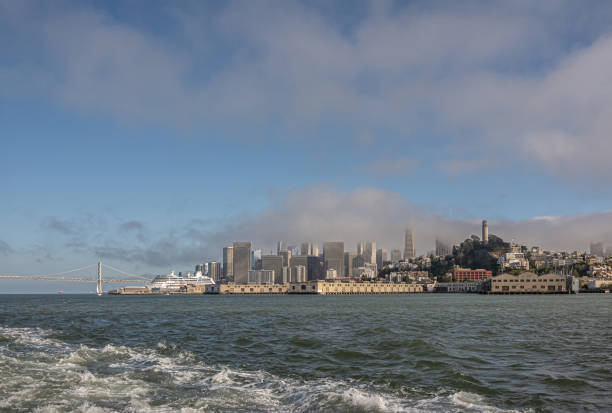 San Francisco, CA, USA - July 12, 2023: financial district behind harbor docks. Cityscape view from gray bay water under blue cloudscape. SF-Oakland bridge and white docked cruise ship