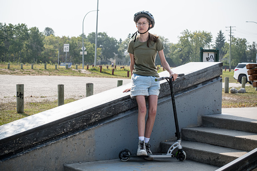 A testament to youthful bravery, this 13-year-old, donning a helmet for safety, leans confidently against the skatepark structure, scooter by her side, as she embarks on her solo adventure through the twists and turns of the park