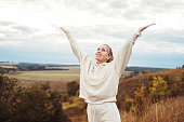 A happy woman in a tracksuit stands on a hilltop on a summer day and enjoys unity with nature, raising her hands to the sky.