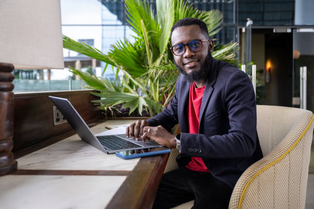 Candid portrait of African professional using laptop Three-quarter front view of man with beard, wearing eyeglasses and casual suit, pausing from typing to smile at camera in lounge of modern office building. kenyan man stock pictures, royalty-free photos & images