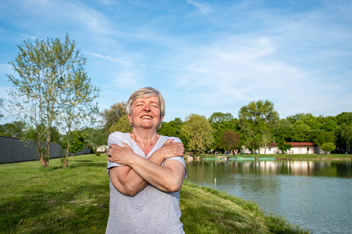 an elderly woman with gray hair stands by the lake and hugs herself