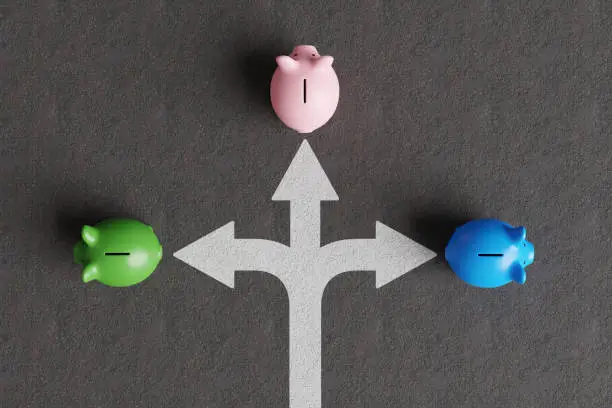 Photo of White divided road sign mark on asphalt with 3 different colored piggy banks going to different directions. Illustration of the concept of stock picks and varied investment products