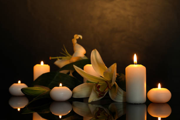 istock White lilies and burning candles on black mirror surface in darkness, space for text. Funeral symbols 1653425826
