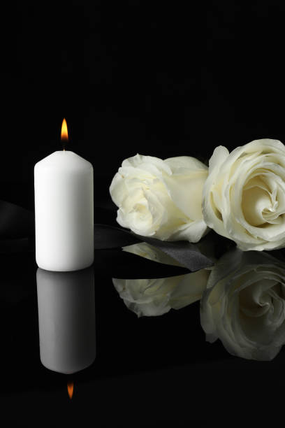 istock Burning candle, white roses and ribbon on black mirror surface in darkness, space for text. Funeral symbols 1653428107