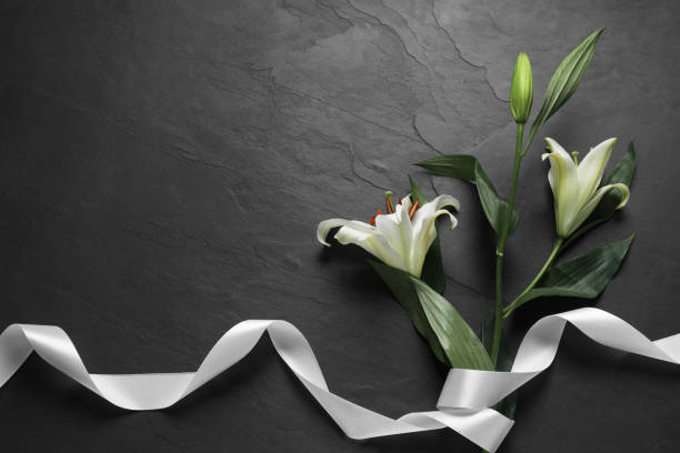 istock White lilies and ribbon on black table, flat lay with space for text. Funeral symbols 1653429069
