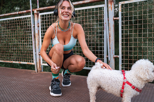Young active Caucasian sportswoman listening to music on wireless headphones while petting dog, on the bridge