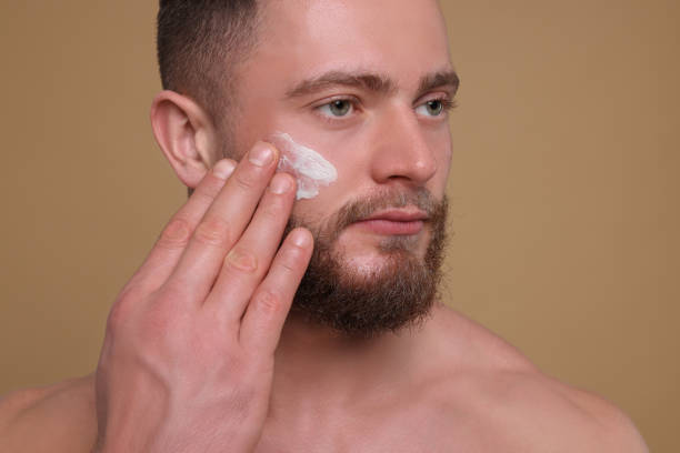 istock Handsome man applying cream onto his face on pale brown background 1653470512