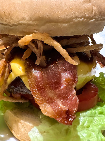 Stock photo showing close-up view of double cheeseburger served in a seedless burger bread bun, an unhealthy and greasy but delicious comfort meal with tomatoes, melted cheese, lettuce, bacon and onion.