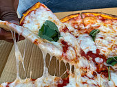 Close-up image slice of Pizza Margherita lifted out of cardboard takeaway pizza box by unrecognisable person, strings of melted golden buffalo mozzarella cheese and rich tomato marinara sauce topping, denim background, elevated view, focus on foreground