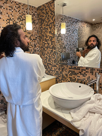 Stock photo showing luxury bathroom, with brown mosaic tiles, white ceramic sink with dual knob monobloc mixer tap on white and grey marble, counter top in front of mirror reflecting an Indian man wearing white towelling robe whilst using a hair dryer to dry his hair after having a shower. Self care and grooming concept.