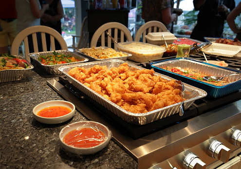 Variety of takeout and homemade food for a family reunion in a Toronto home. Selective focus on deep fried food with dipping sauce on a kitchen island. Family members stand in the background near a window.