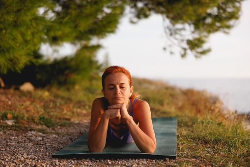 Mature woman in sports clothing lying on front on exercise mat and leaning on elbows with eyes closed,  looking serene and relaxing in nature by the sea, health and body care