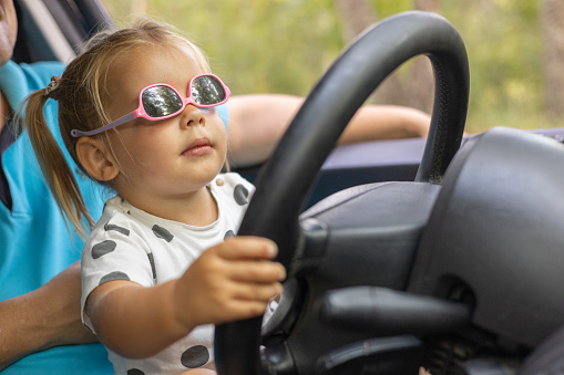 A cute little girl driving a car on her father's lap. Child girl in sunglasses with a serious expression learns to steer a car. A father teaches his daughter how to drive a car..
