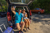 A man and a woman take a selfie while sitting in the open trunk of a car while traveling. People stopped in the forest on the side of the road to take a selfie as a memento of the trip.