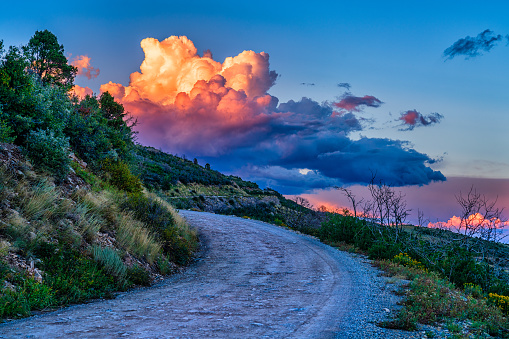 Dramatic Mushroom Cloud Above Mountain Dirt Road S Curve - Scenic view at sunset with road leading off into distance with colorful pink and orange dramatic clouds above in sky. With plenty of copy space and complementary color scheme.