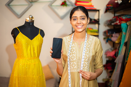 Portrait of Young happy indian woman,tailor,smiling fashion designer showing smartphone screen or display,standing with measurement tap around neck in garment workshop or studio.Mockup concept