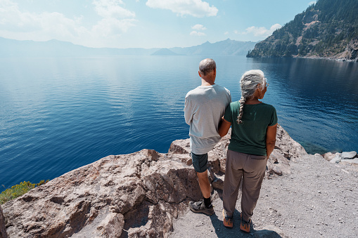 Rear view of a Pacific Islander woman and her Caucasian husband admiring a scenic view of Crater Lake from a cliff edge while on a hike on a warm and sunny summer day.