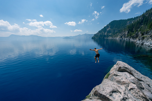 An adventurous and fit man leaps from the rocky cliffside overlooking Crater Lake in Oregon into the frigid and serene waters below on a warm and sunny summer day.