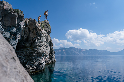 A fit young man and his father-in-law stand on a rocky cliff overlooking Crater Lake in Oregon and ponder jumping in while looking down at the frigid waters below. It is a warm and sunny summer day.