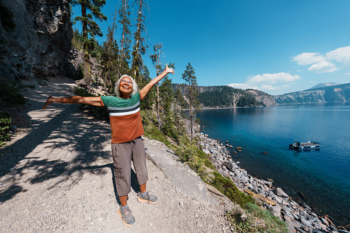 A fit and healthy multiracial senior woman takes a break from hiking to enjoy the scenic view of Crater Lake in Oregon with arms raised to embrace the beauty of nature surrounding her.