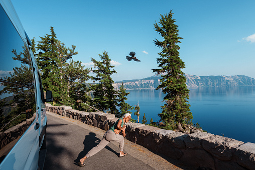 A fit and adventurous senior woman of Hawaiian and Chinese descent takes a break from her retirement roadtrip through the Pacific Northwest region of the United States to practice yoga outside her camper van which is parked at a scenic viewpoint overlooking Crater Lake in Oregon as a bird flies overhead on a beautiful and sunny summer day.