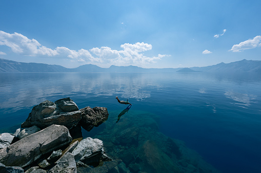 An active and adventurous senior woman of Pacific Islander descent dives off of a rock into the serene and frigid waters of Crater Lake while hiking during a fun camping trip through the Pacific Northwest region of the United States.