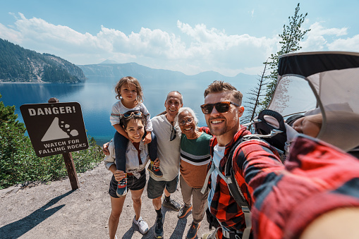An active man who is carrying his one year old son in a backpack baby carrier poses with his wife, toddler daughter, and parents-in-law for a selfie photograph while on a hike overlooking Crater Lake in Oregon on a hot and sunny summer day.