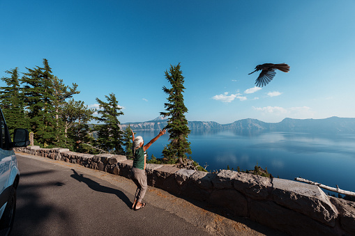 A fit and adventurous senior woman of Hawaiian and Chinese descent takes a break from her retirement roadtrip through the Pacific Northwest region of the United States to practice yoga outside her camper van which is parked at a scenic viewpoint overlooking Crater Lake in Oregon as a bird flies overhead on a beautiful and sunny summer day.