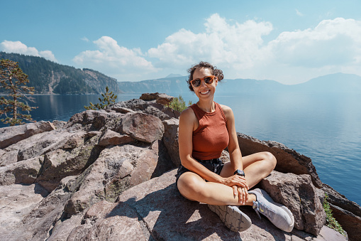 Portrait of  fit Eurasian woman sitting on a rocky cliff edge and smiling directly at the camera while admiring the view of a serene mountain lake on a warm and sunny summer afternoon.