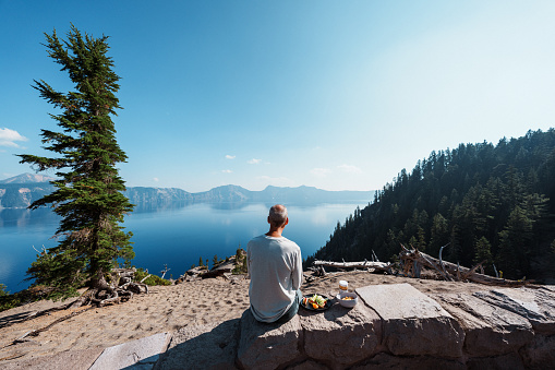 Rear view of a fit senior man sitting on a rock wall and eating lunch while admiring the serene and scenic view of Crater Lake Nation Park on a sunny summer day in Oregon.