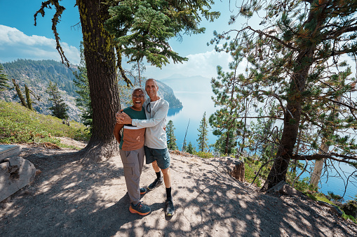 An affectionate and healthy multiracial senior couple takes a break from hiking to pose and smile directly at the camera at a scenic viewpoint overlooking Crater Lake in Oregon during a hike while on vacation.