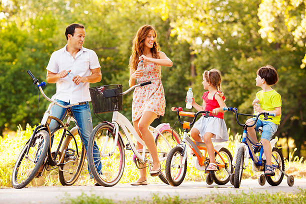 A family of four riding bikes in a park. A man is helping a child on a bike