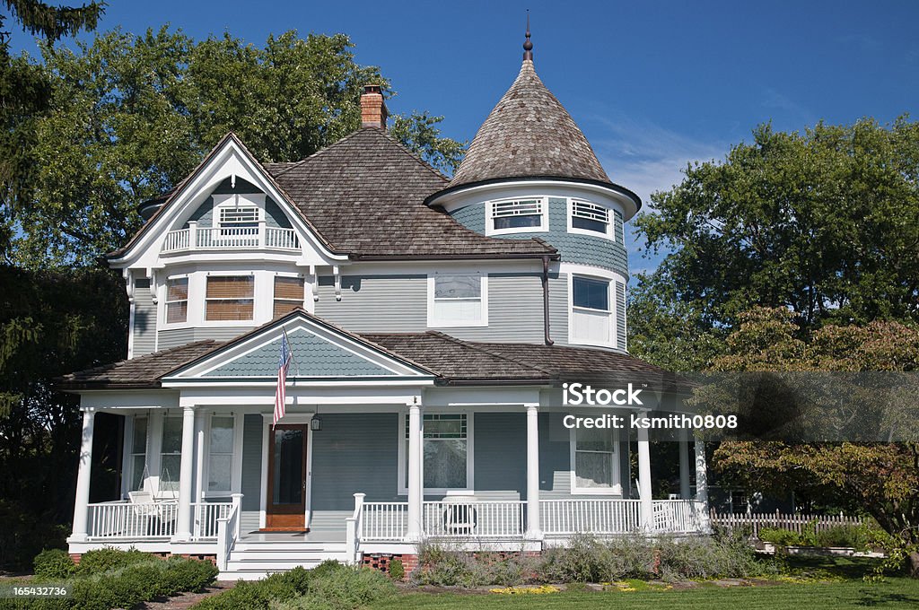 A Victorian style home with wrap-around porch Beautiful gray traditional victorian house.  House has an American Flag haning over the porch and shows a beautiful garden with flowers and trees.  Set against a cloudless blue sky House Stock Photo