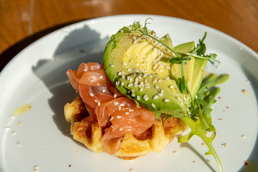 Close-up view of potato waffle with fresh lightly salted salmon, green avocado slices, salad leafs and sesame seeds on white plate on wooden table in restaurant. Morning light. Healthy breakfast theme
